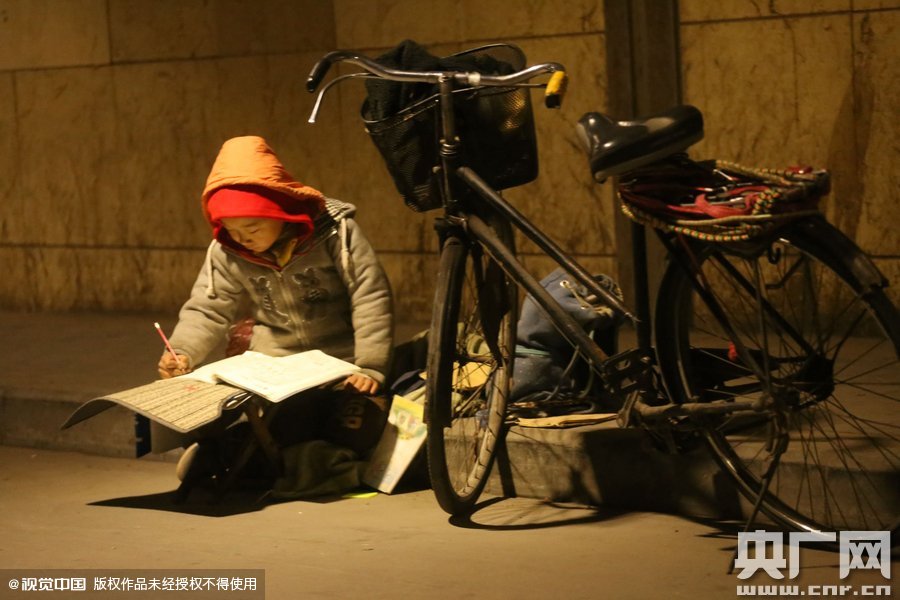 鄭州中原路京廣鐵路大橋下涵洞的馬路牙子邊，一位戴著紅色辣椒帽的小男孩趴在一個小馬扎上，上面鋪著一張車墊，在微弱的燈光下寫作業(yè)