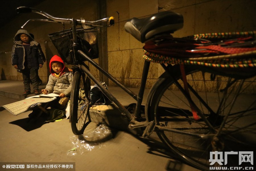 2015年11月8日晚，鄭州，7歲男孩涵洞橋下陪父母練攤，路邊燈下坐小馬扎上寫作業(yè)。